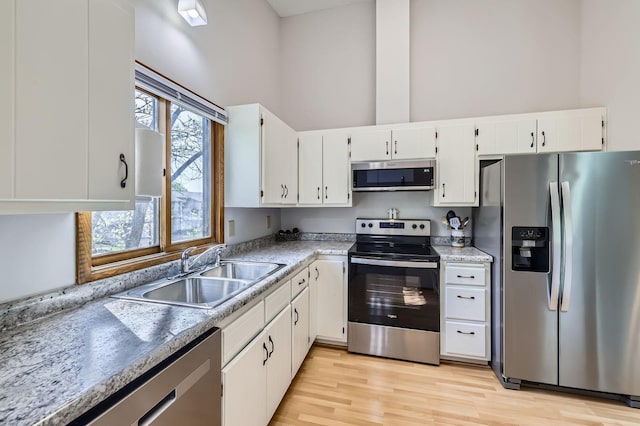 kitchen with appliances with stainless steel finishes, a sink, a towering ceiling, white cabinetry, and light wood-type flooring