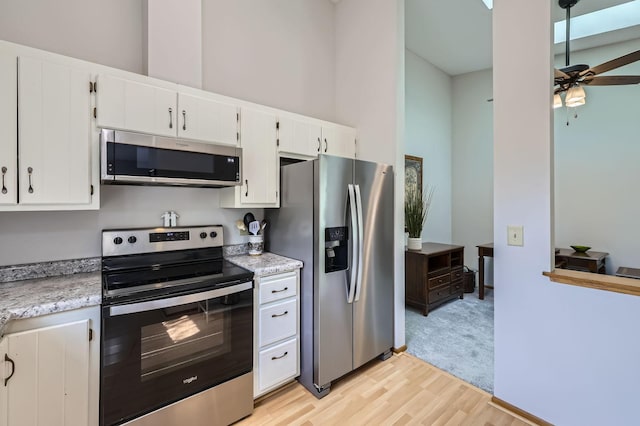 kitchen featuring appliances with stainless steel finishes, ceiling fan, light wood-type flooring, white cabinetry, and a towering ceiling