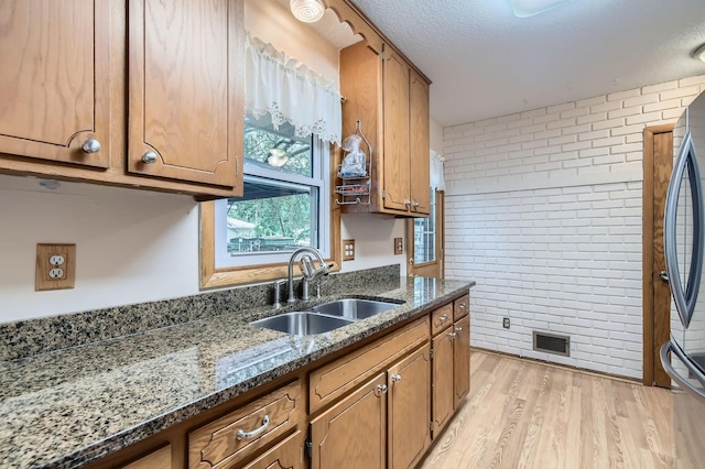 kitchen featuring a sink, brick wall, brown cabinets, light wood-style flooring, and dark stone countertops