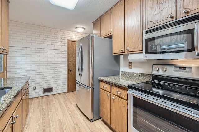kitchen with appliances with stainless steel finishes, brick wall, a textured ceiling, light wood-style floors, and light stone countertops