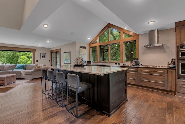 27806 Sundance Ln, Cold Spring, MN 56320 - kitchen featuring wall chimney exhaust hood, a kitchen island, a breakfast bar area, light stone countertops, and dark wood-type flooring