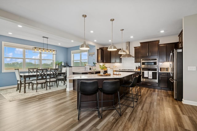 19281 Blue Stem Ct, Maple Grove, MN 55311 - kitchen with wall chimney exhaust hood, appliances with stainless steel finishes, dark wood-style floors, light countertops, and dark brown cabinets
