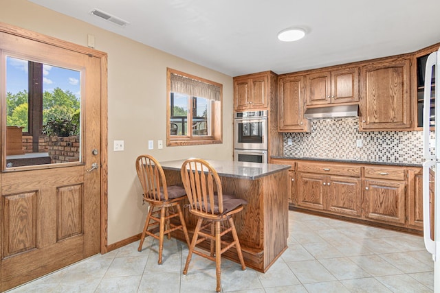 17000 Jackal St NW, Ramsey, MN 55303 - kitchen with under cabinet range hood, stainless steel double oven, brown cabinets, tasteful backsplash, and light tile patterned flooring
