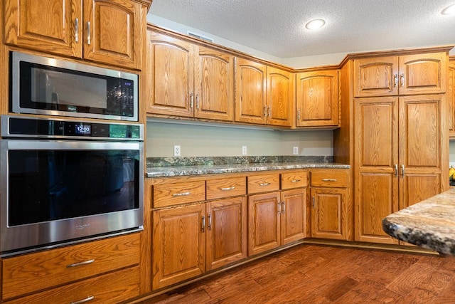 18505 210th Ave NE, Paynesville, MN 56362 - kitchen with appliances with stainless steel finishes, a textured ceiling, brown cabinets, dark wood-style flooring, and stone countertops