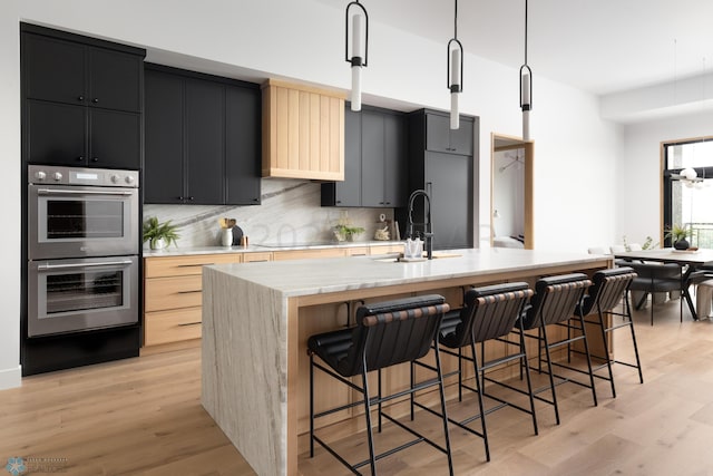 kitchen featuring light brown cabinets, a center island with sink, double oven, decorative light fixtures, and a kitchen bar