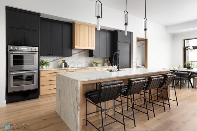 kitchen with hanging light fixtures, backsplash, double oven, a kitchen bar, and a kitchen island with sink