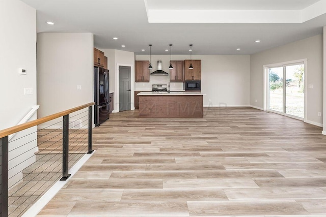 kitchen featuring light wood-type flooring, wall chimney range hood, an island with sink, black appliances, and hanging light fixtures