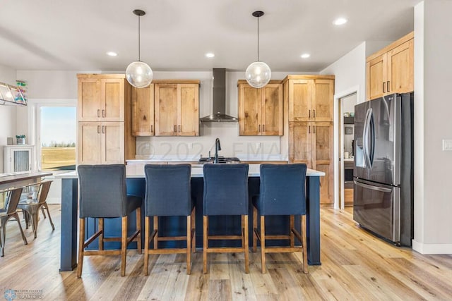 kitchen with wall chimney range hood, a center island with sink, light countertops, light wood-style floors, and stainless steel fridge
