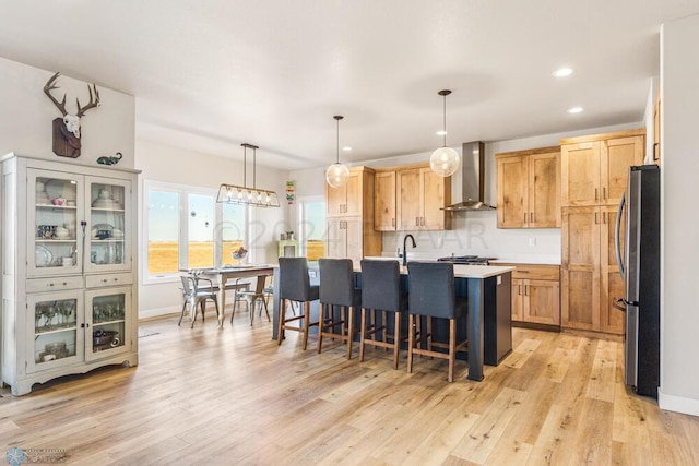 kitchen featuring a center island with sink, a kitchen bar, light wood-style flooring, freestanding refrigerator, and wall chimney exhaust hood