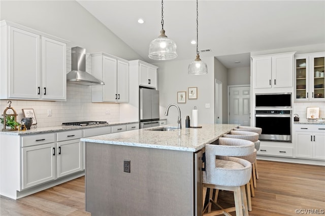 kitchen with appliances with stainless steel finishes, wall chimney exhaust hood, white cabinetry, a kitchen island with sink, and light wood-type flooring