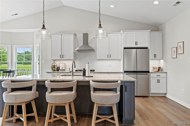 kitchen with stainless steel refrigerator, lofted ceiling, a kitchen island with sink, and wall chimney exhaust hood