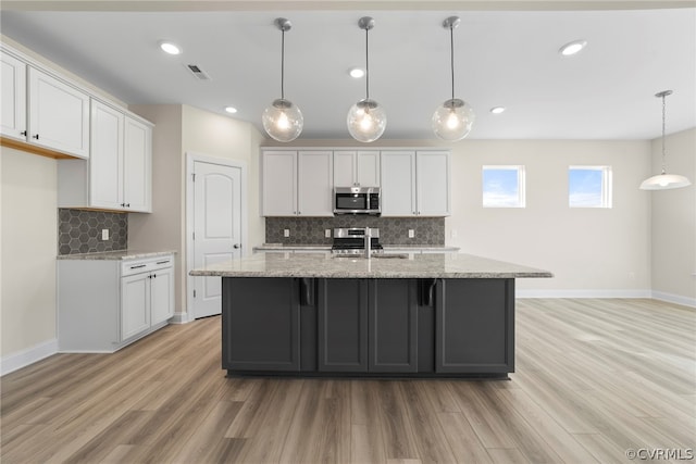 kitchen featuring pendant lighting, stainless steel appliances, light stone countertops, an island with sink, and white cabinets