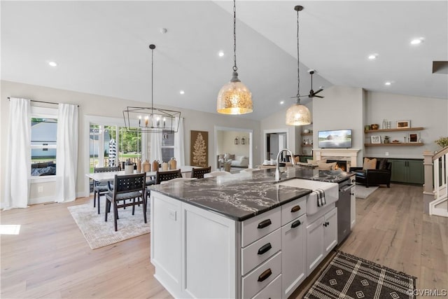 kitchen with a center island with sink, dark stone counters, light wood-type flooring, a warm lit fireplace, and a sink