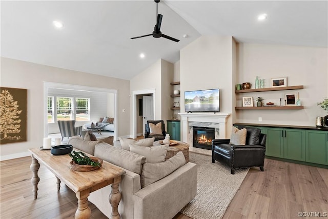 living area featuring baseboards, ceiling fan, light wood-type flooring, a premium fireplace, and high vaulted ceiling