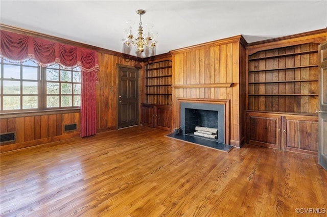 unfurnished living room with built in features, a fireplace, wood walls, hardwood / wood-style flooring, and a notable chandelier