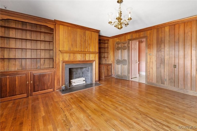 unfurnished living room with a notable chandelier, built in shelves, wooden walls, and light hardwood / wood-style floors