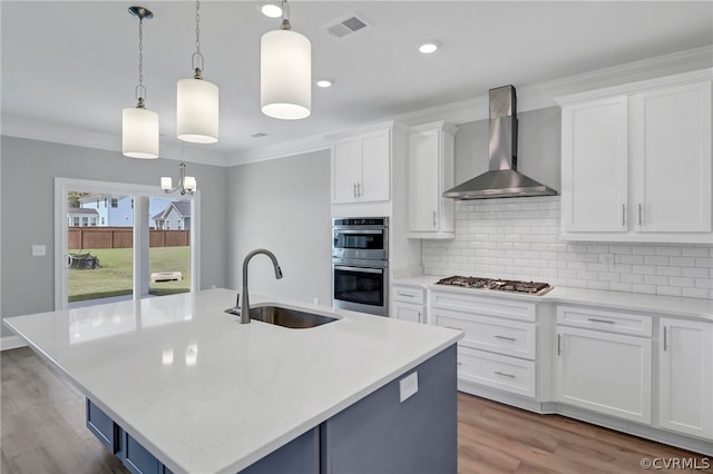 kitchen with wall chimney range hood, hanging light fixtures, appliances with stainless steel finishes, a kitchen island with sink, and sink