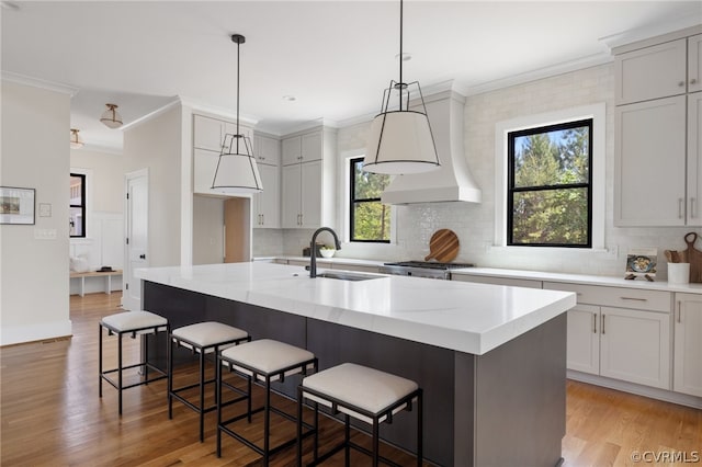 kitchen with custom range hood, light hardwood / wood-style flooring, a kitchen island with sink, light stone counters, and decorative light fixtures