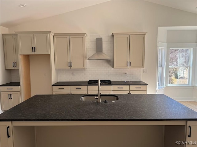 kitchen with backsplash, a kitchen island with sink, vaulted ceiling, wall chimney range hood, and a sink