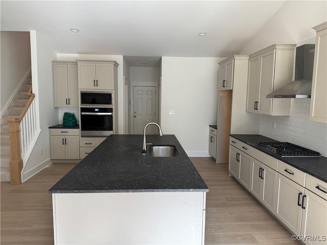 kitchen with light wood finished floors, appliances with stainless steel finishes, a kitchen island with sink, a sink, and wall chimney exhaust hood