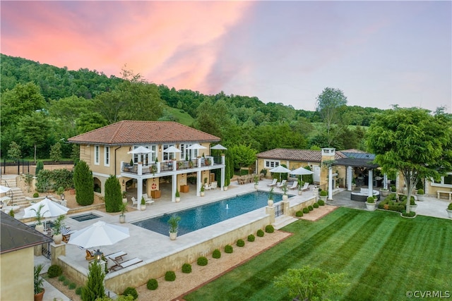 back house at dusk featuring a balcony, a patio area, a yard, a fenced in pool, and a pergola