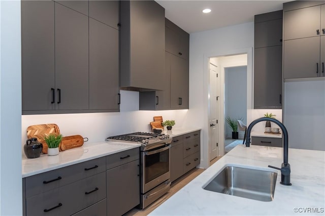 kitchen featuring stainless steel gas range oven, gray cabinetry, light stone counters, wall chimney exhaust hood, and a sink