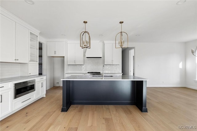 kitchen with stainless steel microwave, hanging light fixtures, premium range hood, a kitchen island with sink, and light wood-type flooring