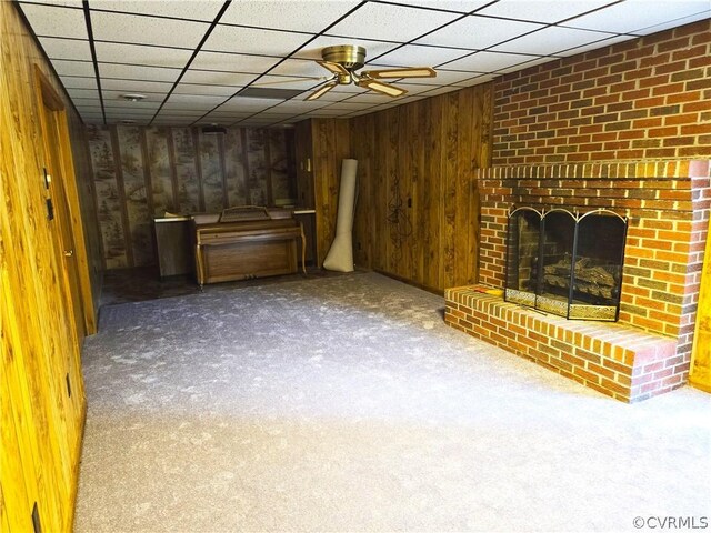 unfurnished living room with a drop ceiling, carpet flooring, wooden walls, ceiling fan, and a fireplace