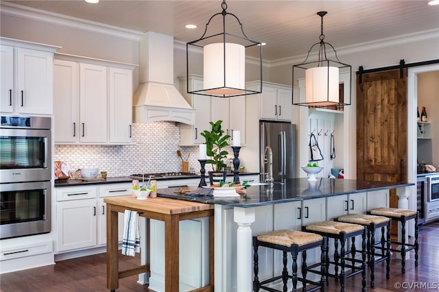 kitchen featuring dark wood-type flooring, custom range hood, appliances with stainless steel finishes, a kitchen island with sink, and a barn door