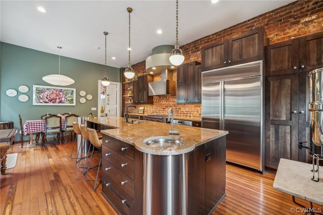 kitchen featuring appliances with stainless steel finishes, light wood-type flooring, a center island with sink, and wall chimney exhaust hood