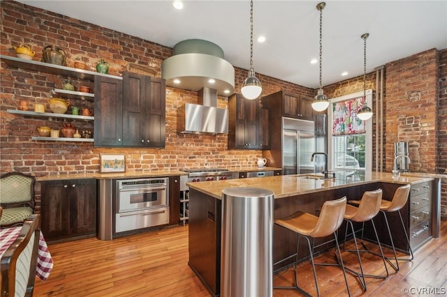 kitchen with dark brown cabinetry, wall chimney range hood, pendant lighting, a center island with sink, and appliances with stainless steel finishes