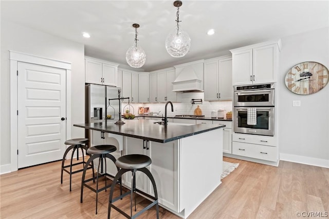 kitchen featuring white cabinetry, stainless steel appliances, premium range hood, light hardwood / wood-style floors, and a kitchen island with sink