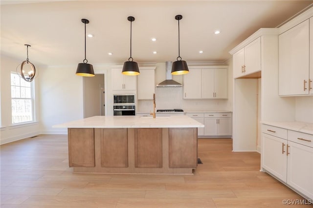 kitchen featuring pendant lighting, wall chimney range hood, light stone counters, an island with sink, and white cabinets