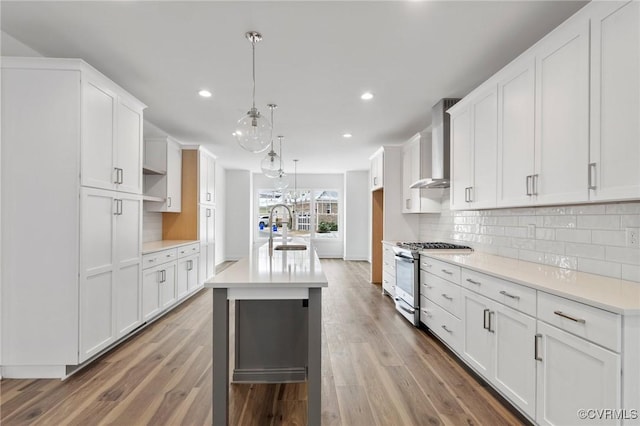 kitchen with stainless steel range with gas stovetop, wall chimney range hood, decorative light fixtures, white cabinetry, and a kitchen island with sink