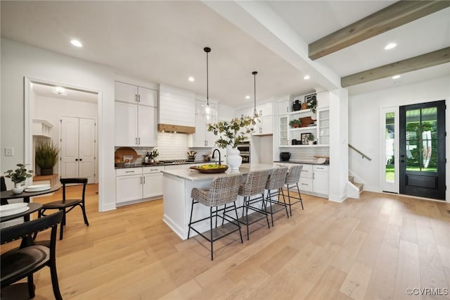 kitchen featuring white cabinetry, a kitchen island with sink, light hardwood / wood-style floors, decorative light fixtures, and beamed ceiling