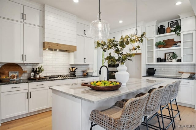 kitchen featuring dark stone countertops, hanging light fixtures, an island with sink, and white cabinets