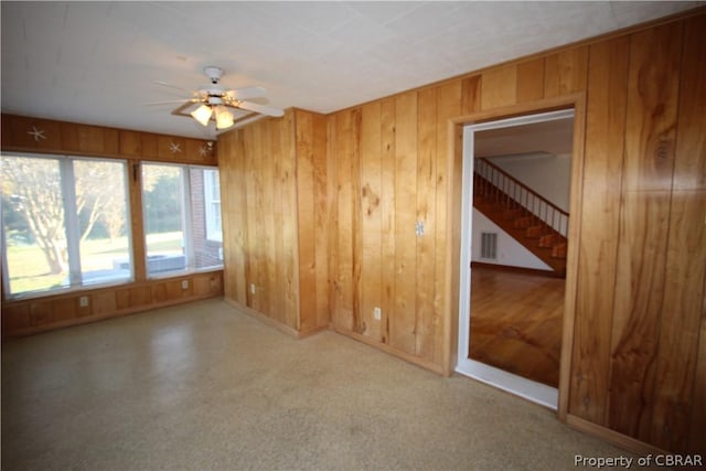 unfurnished room featuring wooden walls, baseboards, visible vents, a ceiling fan, and stairway