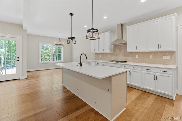 kitchen featuring decorative light fixtures, white cabinets, wall chimney range hood, a center island with sink, and light wood-type flooring