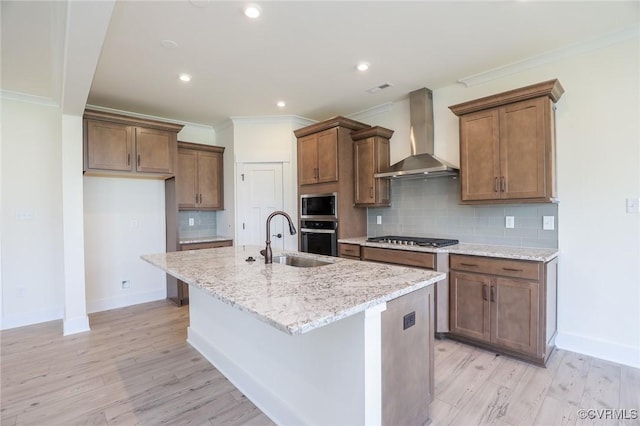 kitchen featuring wall chimney exhaust hood, a sink, appliances with stainless steel finishes, a center island with sink, and light wood finished floors