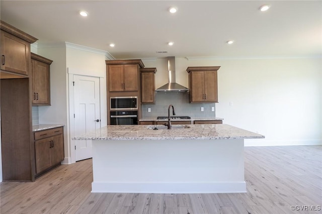 kitchen featuring wall chimney exhaust hood, stainless steel appliances, ornamental molding, an island with sink, and light stone countertops