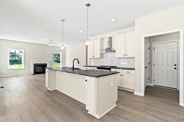 kitchen with wall chimney range hood, tasteful backsplash, pendant lighting, a kitchen island with sink, and white cabinets