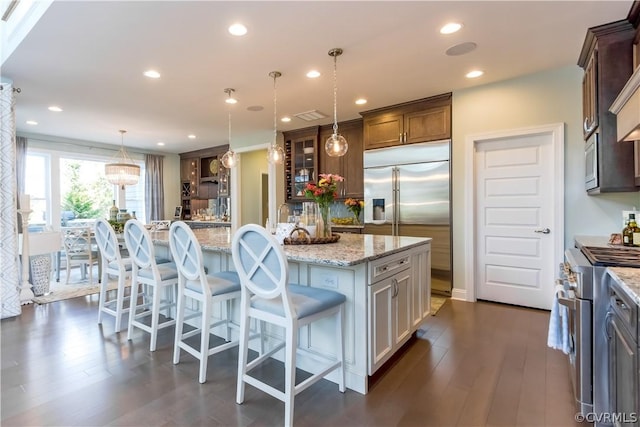 kitchen with a spacious island, hanging light fixtures, built in appliances, light stone countertops, and a kitchen bar