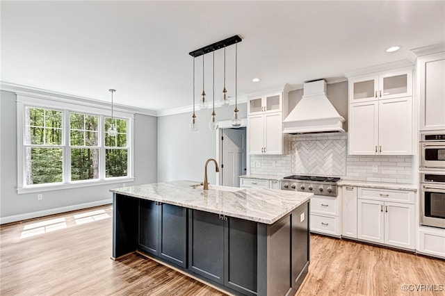 kitchen with premium range hood, sink, stainless steel gas cooktop, light hardwood / wood-style flooring, and a center island with sink