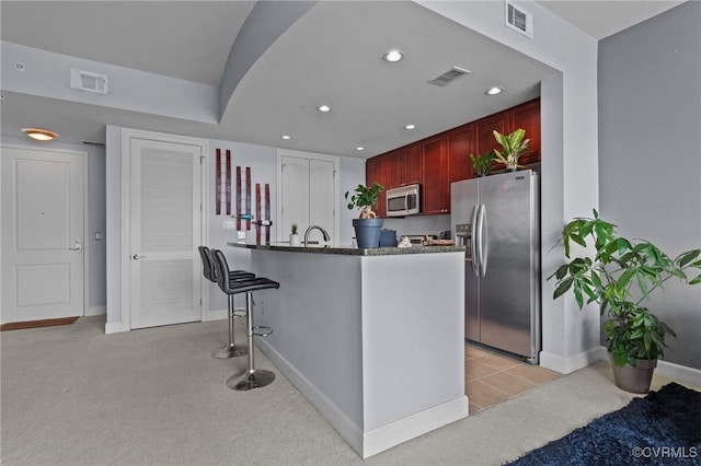 kitchen featuring dark stone countertops, kitchen peninsula, appliances with stainless steel finishes, light carpet, and a kitchen bar