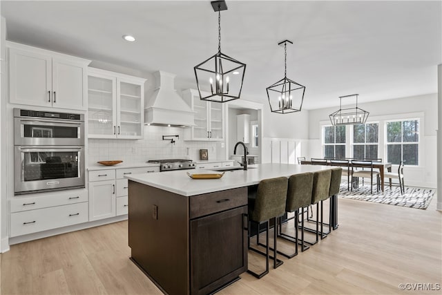 kitchen with light wood-type flooring, stainless steel double oven, decorative light fixtures, and premium range hood