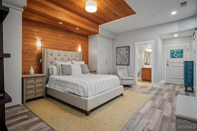 bedroom with connected bathroom, light wood-type flooring, and wooden walls