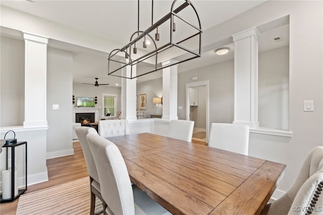 dining area featuring a ceiling fan, baseboards, decorative columns, a lit fireplace, and light wood-type flooring