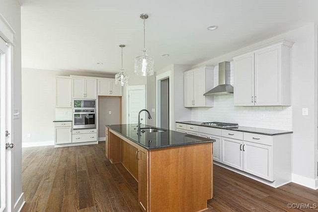 kitchen featuring dark countertops, dark wood finished floors, appliances with stainless steel finishes, wall chimney exhaust hood, and a sink