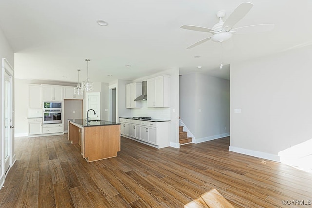 kitchen with wall chimney range hood, ceiling fan, an island with sink, and white cabinetry