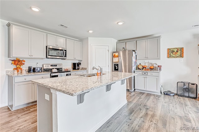 kitchen with light wood-type flooring, a kitchen island with sink, stainless steel appliances, light stone counters, and sink